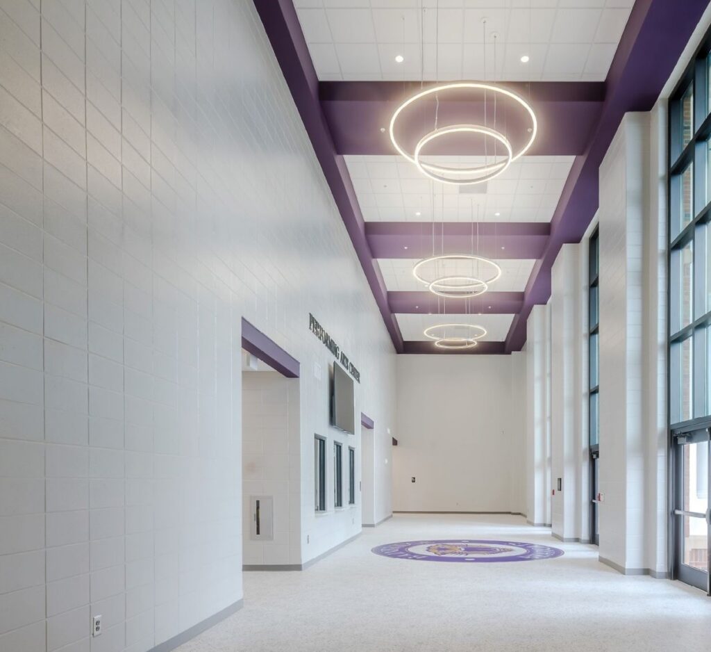A spacious modern hallway with white walls, purple ceiling accents, circular light fixtures, tall windows, and a round floor emblem.