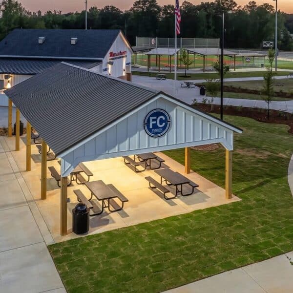 Illuminated pavilion with picnic tables near a concession stand and ball fields at sunset, with walkways, lawn, and playground nearby.