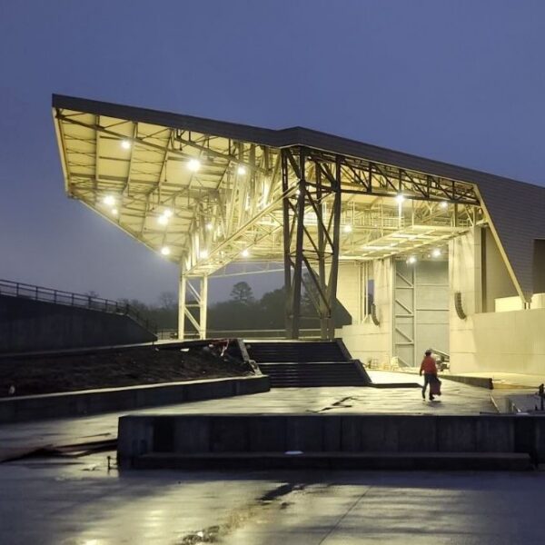 An outdoor amphitheater with a large metal roof glows at dusk. Wet ground reflects lights. A person in a red jacket nearby.