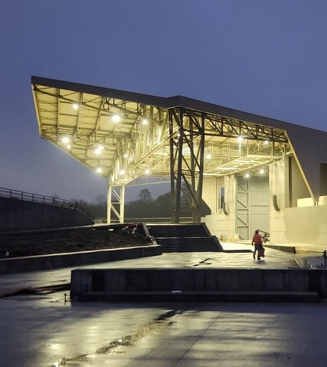 An outdoor amphitheater with a large metal roof glows at dusk. Wet ground reflects lights. A person in a red jacket nearby.