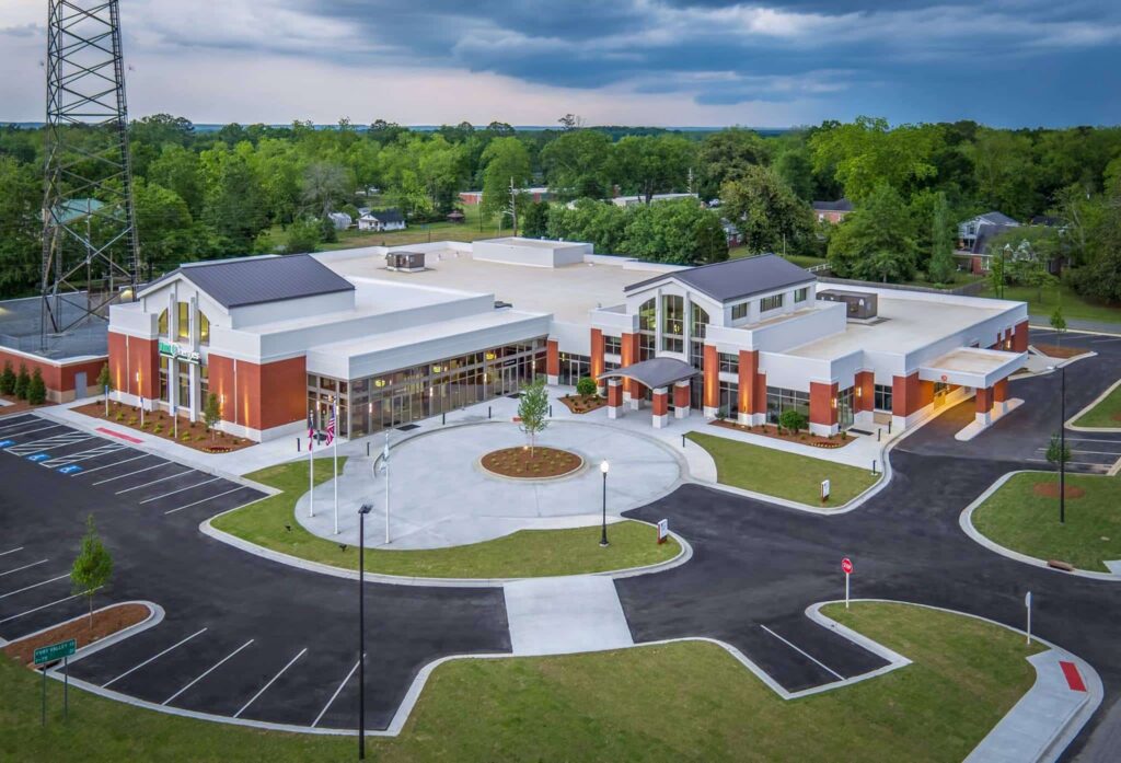 Aerial view of a modern single-story brick building featuring a circular driveway, adjacent parking lots, and neatly landscaped grounds, all surrounded by trees beneath a partly cloudy sky.