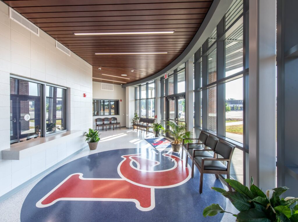 A bright, modern lobby featuring expansive windows, a wooden ceiling, chairs, and lush plants. A large red and blue school logo decorates the floor as sunlight fills the space, enhancing its welcoming atmosphere.