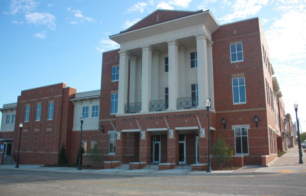 A three-story red brick building with white columns at the entrance, labeled Batte College Center. The sky is clear and blue, and the street in front is empty.