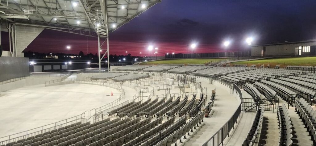 Empty outdoor amphitheater with rows of seats illuminated by stadium lights, curving around a concrete stage. The sky is dark, filled with shades of purple and pink from the setting sun in the background.