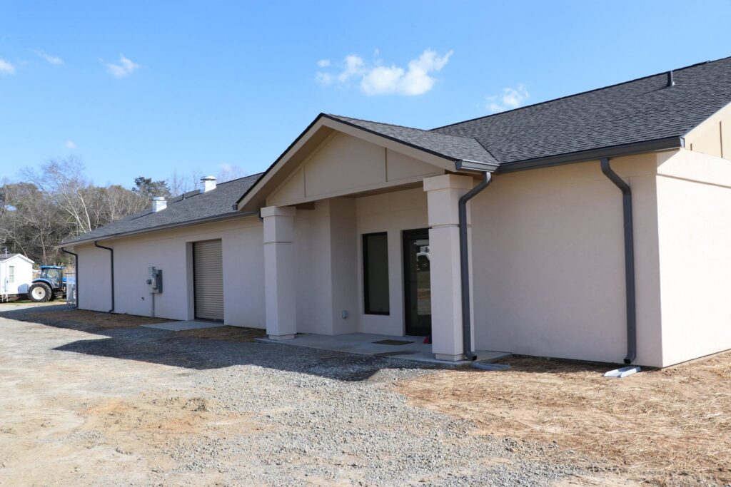 Single-story beige house with dark shingles, a covered entryway, large window, garage door, and visible gutters. The yard is unfinished with dirt and gravel, with trees and a blue sky in the background.