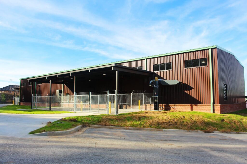 A spacious brown industrial warehouse with a green roof, surrounded by chain-link fencing and featuring open loading bays, stands beneath a clear blue sky on a sunny day.