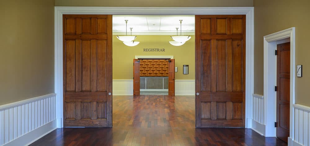 View through open wooden doors into a hallway with wood floors, pale walls, and a sign reading Registrar above a service counter. Ceiling lights hang above, and small side doors are visible on each side.