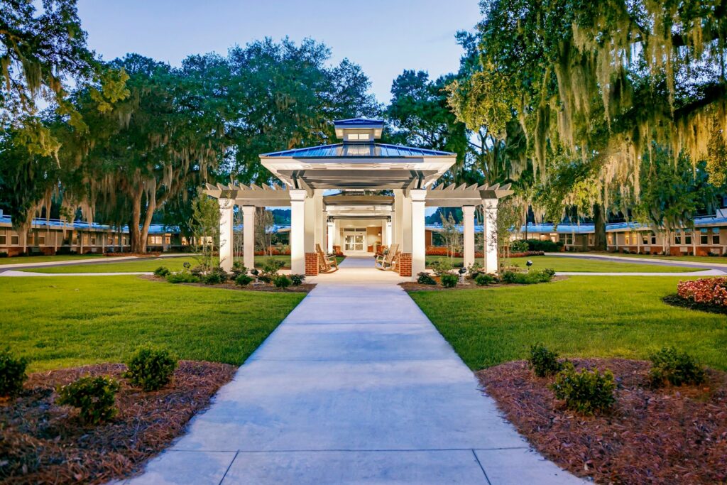A covered outdoor pavilion with white columns stands in the middle of a landscaped courtyard, encircled by green grass, trees draped with hanging moss, and walkways connecting to nearby buildings.