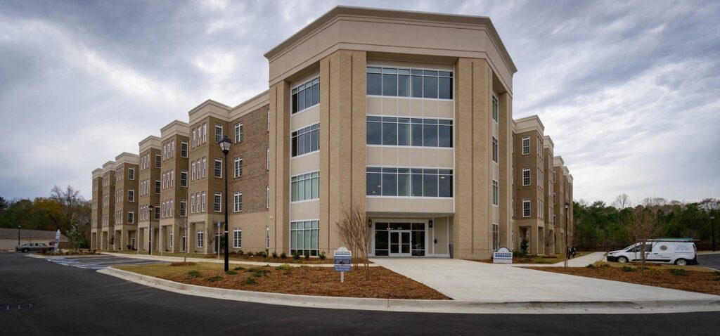 A contemporary three-story brick building featuring large windows, a prominent main entrance, and neatly landscaped grounds beneath a cloudy sky. Near the entrance, a white van and multiple signs can be seen.