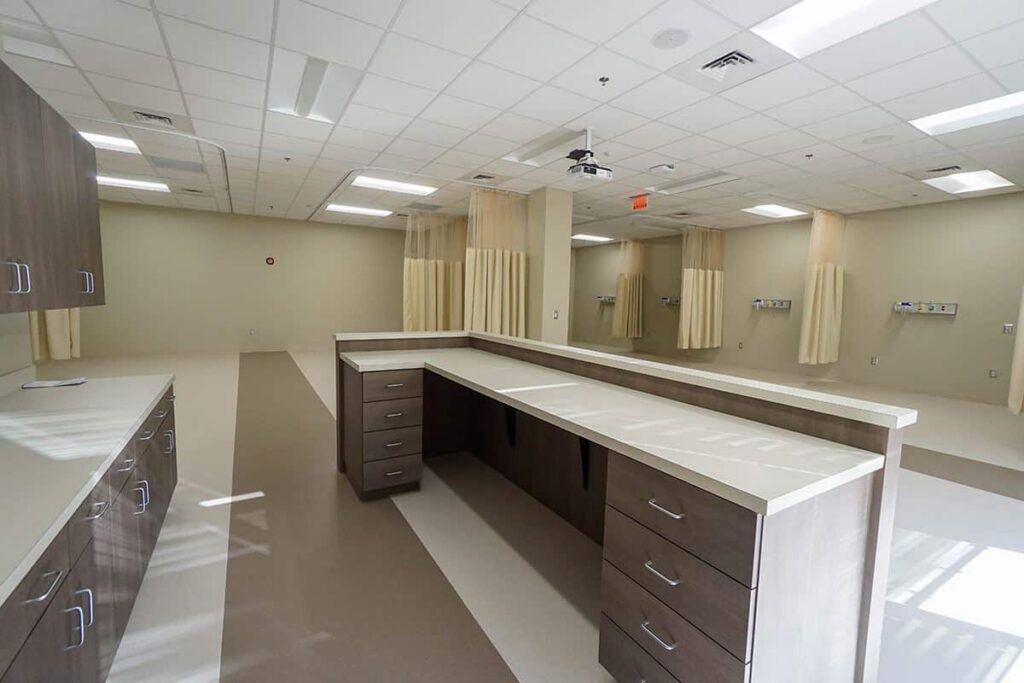 A modern, empty hospital nurse station featuring brown cabinets and cream-colored walls. In the background, several patient areas with curtains are visible beneath bright ceiling lights.