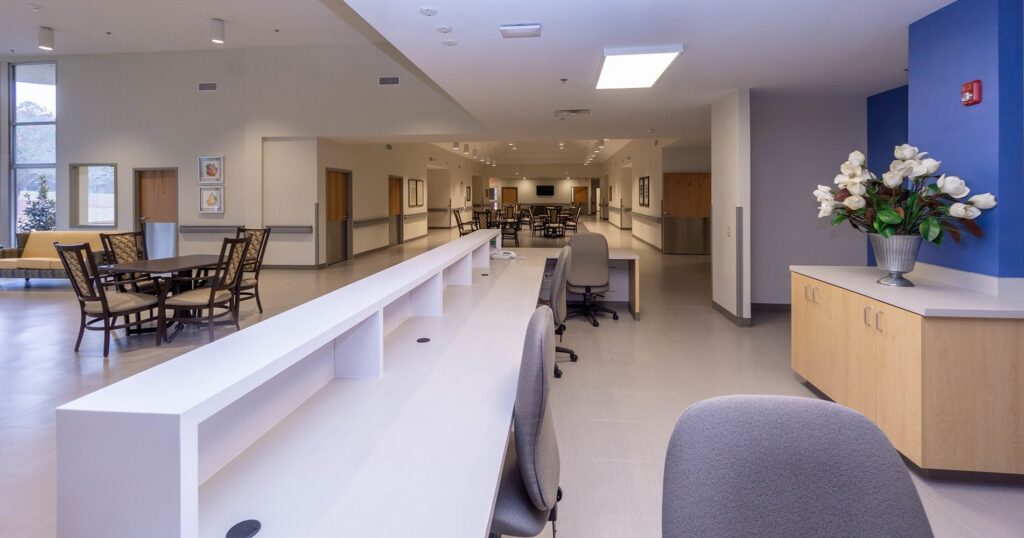 Spacious, modern office or healthcare facility with white desks, gray chairs, light wood cabinets, a blue accent wall, and a decorative vase of white flowers. Several tables and chairs are visible in the background.