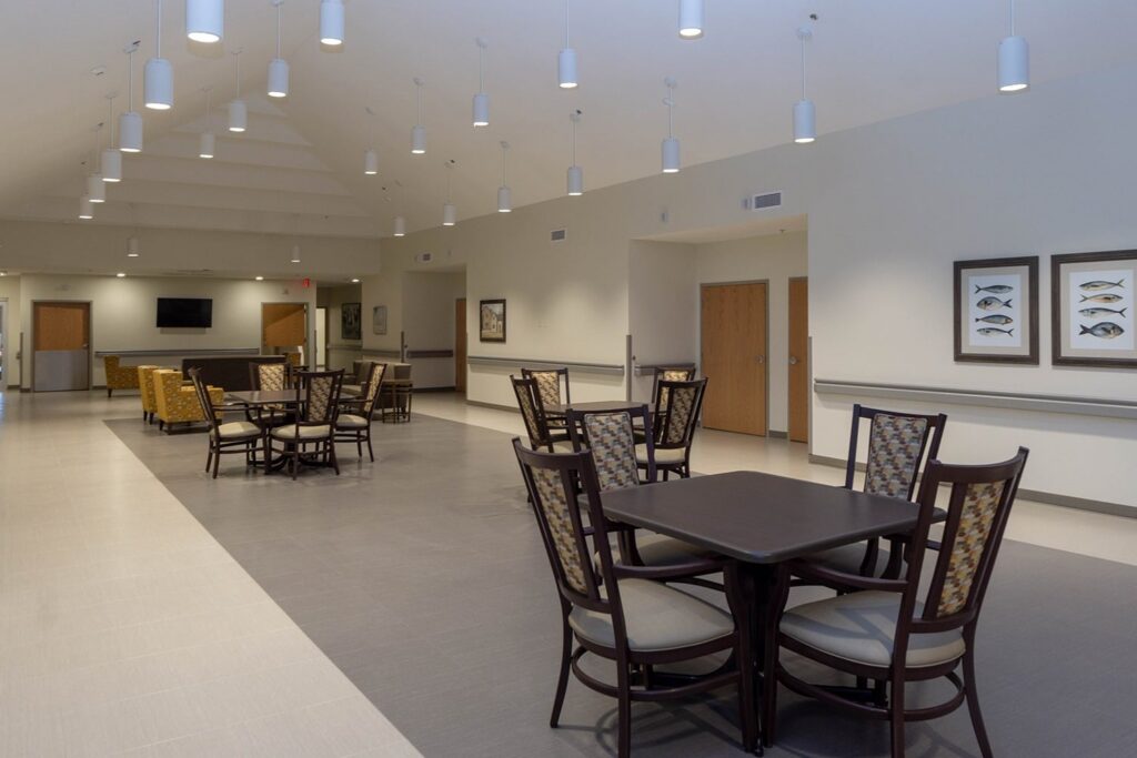 A bright, open room featuring multiple square tables with chairs, neutral-colored walls decorated with framed fish art, and pendant lights hanging from the ceiling, creating a contemporary common or dining area atmosphere.