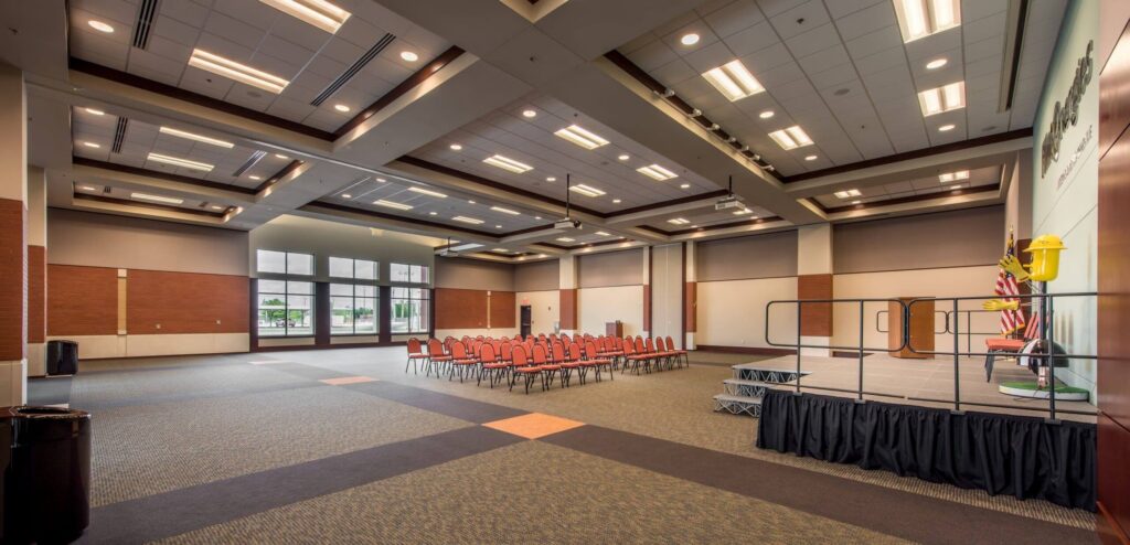 A spacious, empty conference room features rows of red chairs facing a stage. Large windows allow natural light to flood in, and U.S. flags stand at the edge of the stage. Overhead lights brighten the room.