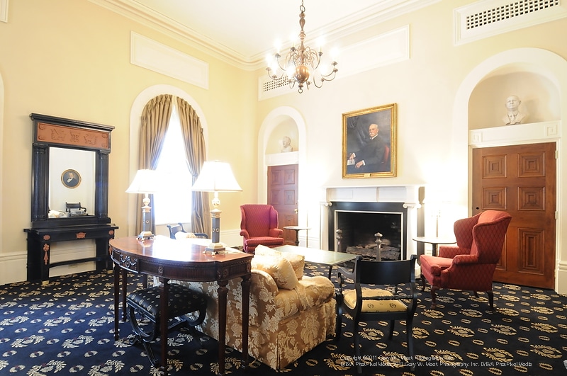 Sophisticated room featuring an ornate rug, vintage furnishings, a chandelier, fireplace beneath a portrait, two red armchairs, a round desk, and expansive windows adorned with gold curtains letting in natural light.