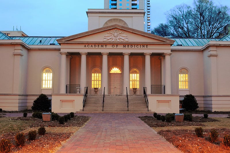 A neoclassical building with columns, labeled Academy of Medicine, lit warmly from inside, with a brick path, bushes, and trees in the foreground and a tall building in the background.
