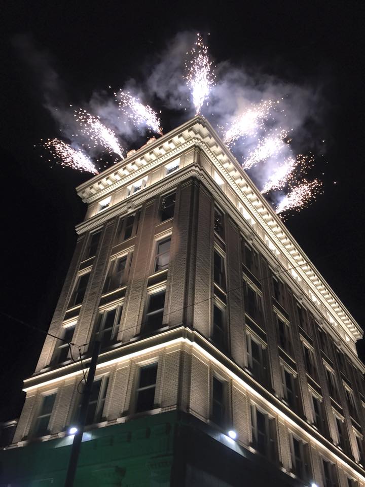 Fireworks burst from the rooftop of a tall, illuminated brick building at night, with smoke trailing into the dark sky above the structure.