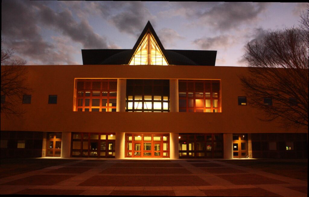 A modern building featuring expansive windows and a triangular glass roof is warmly illuminated at dusk, flanked by bare trees with a cloudy sky overhead.