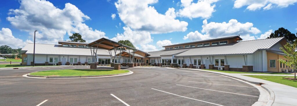 A sleek, single-story building with a metal roof, expansive windows, and a sheltered entrance stands beneath a blue sky dotted with white clouds. The property features a circular driveway and several vacant parking spots.