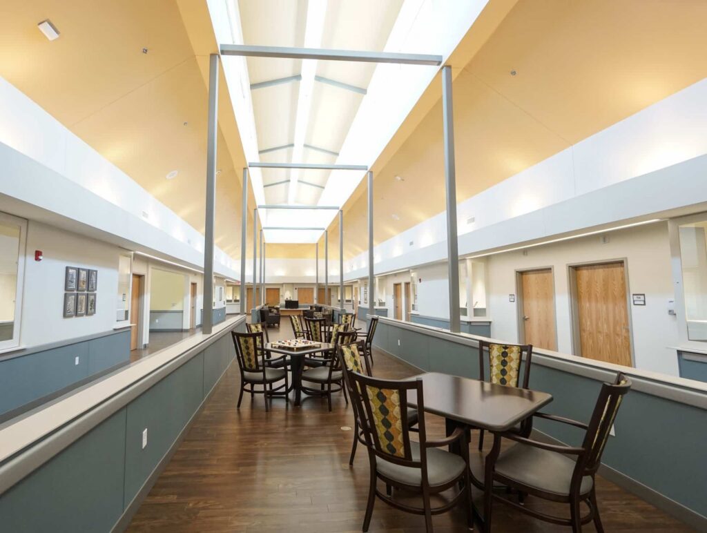 A bright, spacious hallway features wooden floors with tables and chairs neatly arranged for seating. Skylights overhead fill the area with natural light, and multiple doorways are visible along both sides of the corridor.