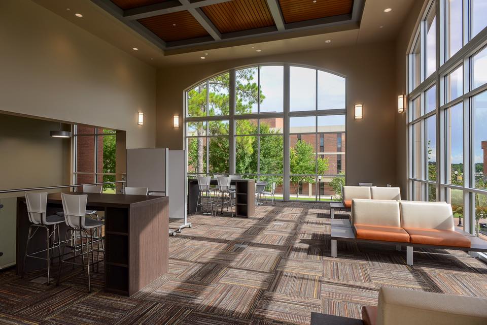 Modern lounge with large windows, sofas, high tables, and chairs. Sunlight streams in, creating patterns on the carpet. Trees and a brick building are visible outside. The space feels bright and inviting.
