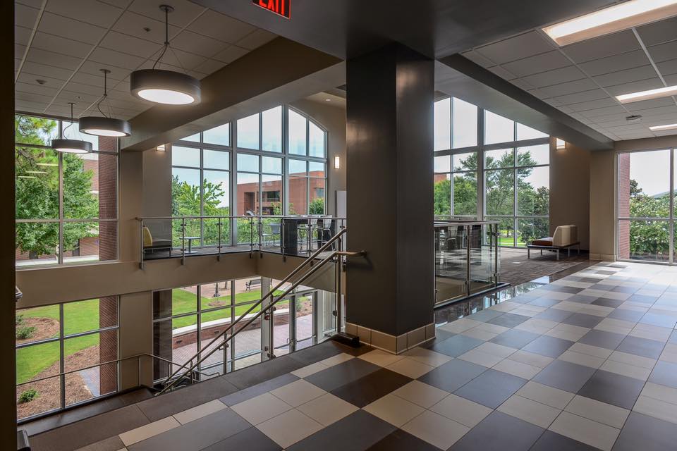 Modern building interior with large windows, tile flooring, metal railings, and a staircase. Natural light fills the space, showing green trees and a brick building outside.