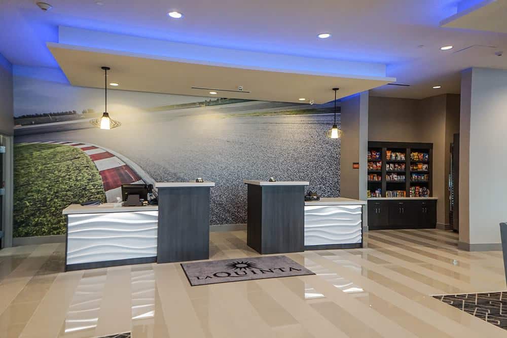 Modern hotel lobby featuring two check-in desks, a racetrack mural on the wall, modern lighting fixtures, and a snack area to the right. A La Quinta-branded rug lies on the striped tile floor.