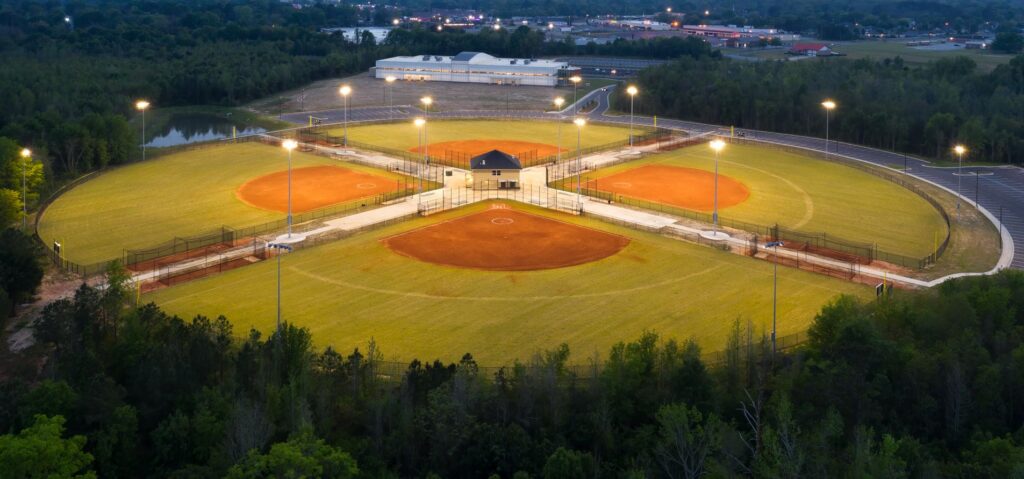 Aerial view of a brightly illuminated baseball complex at dusk, showcasing four neighboring fields laid out in a wagon-wheel pattern, with trees and parking lots encircling the area.