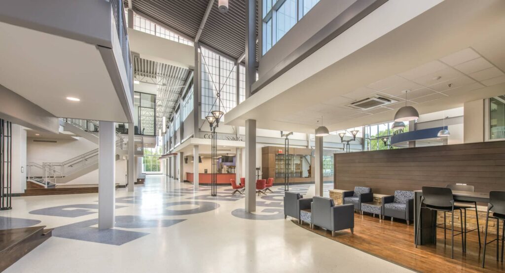 Modern building interior featuring high ceilings and expansive windows, with lounge chairs, tables, a reception desk, and an open staircase. The spacious area is filled with natural light.