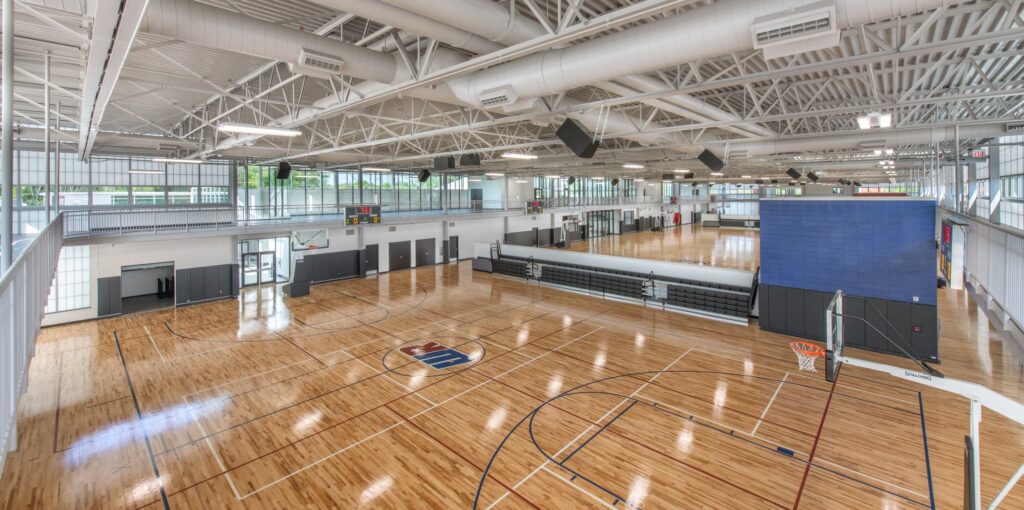 Spacious indoor gymnasium featuring several basketball courts, rows of bleachers, large windows letting in natural sunlight, and exposed white ceiling beams. A prominent logo is displayed at the center of the polished court floor.