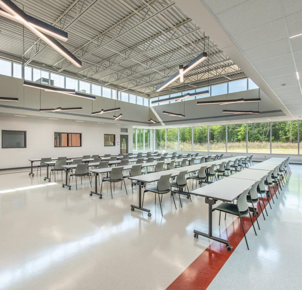 A spacious modern cafeteria features rows of gray tables and chairs, large windows that allow natural light to pour in, and a picturesque view of trees outside. Linear lights hang from the high ceiling above.