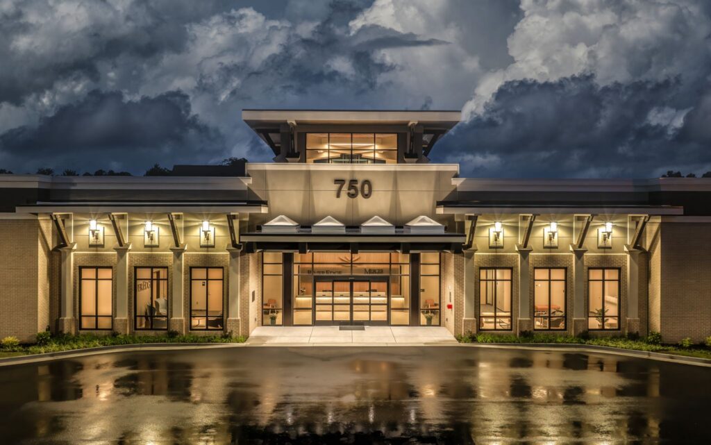 A contemporary building featuring expansive windows and prominent outdoor lighting, with the number 750 displayed above the entrance. The scene is illuminated at dusk, set against a moody sky filled with dramatic clouds and reflections shimmering on the wet pavement below.