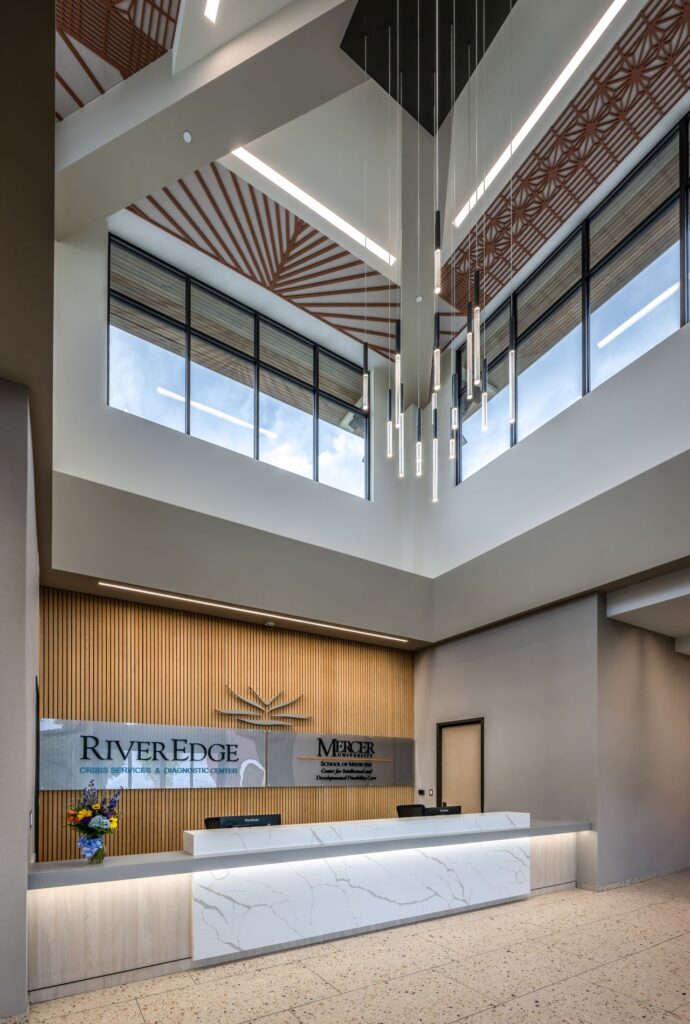 A modern office lobby with a white marble reception desk, pendant lights, tall windows, and wooden wall accents. River Edge and Merzar logos are displayed behind the desk. A small flower arrangement sits on the counter.