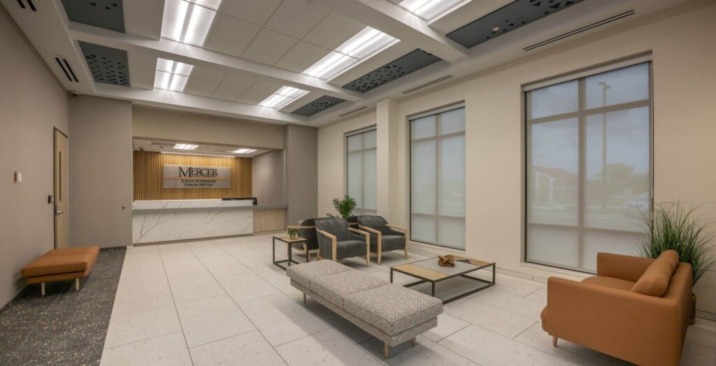 Modern office waiting area with tan and gray seating, a coffee table, potted plants, and large windows. A reception desk with a Mercer sign is at the back of the room under bright ceiling lights.
