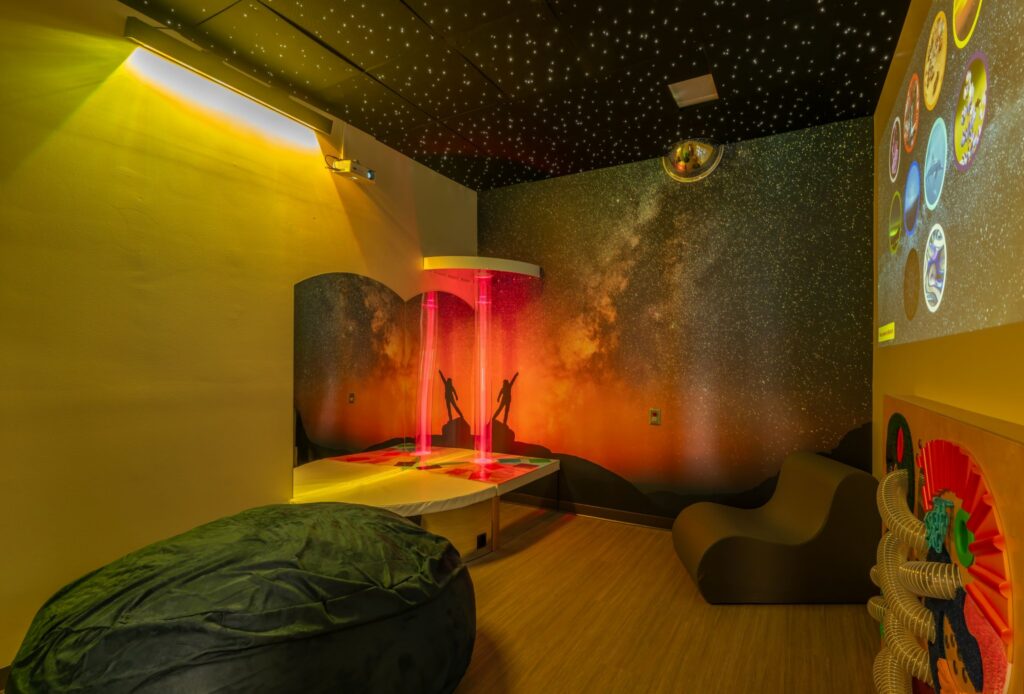 A sensory room featuring a bean bag, soft seating, a water bubble tube glowing with red lights, starry ceiling lights, a galaxy-themed mural on the wall, and various tactile wall activities.
