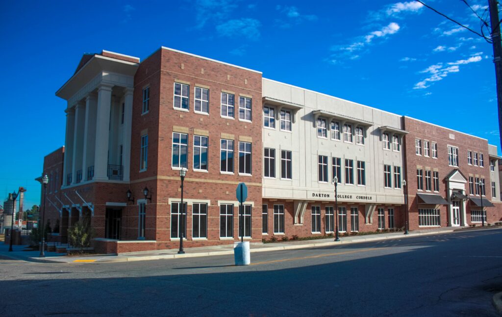 Three-story brick building with tall columns, large windows, and a sign reading Dalton College Center on its facade, set against a bright blue sky with some clouds. The street in front is empty.