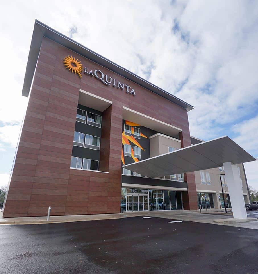 A modern LaQuinta hotel building featuring a large geometric entrance canopy, multiple windows, and the LaQuinta logo displayed on the upper facade. Partly cloudy skies can be seen above the structure.