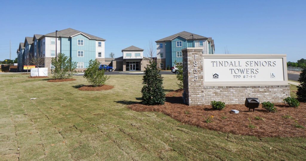 A sign reading Tindall Seniors Towers stands in front of a landscaped lawn and modern apartment buildings under a clear blue sky.