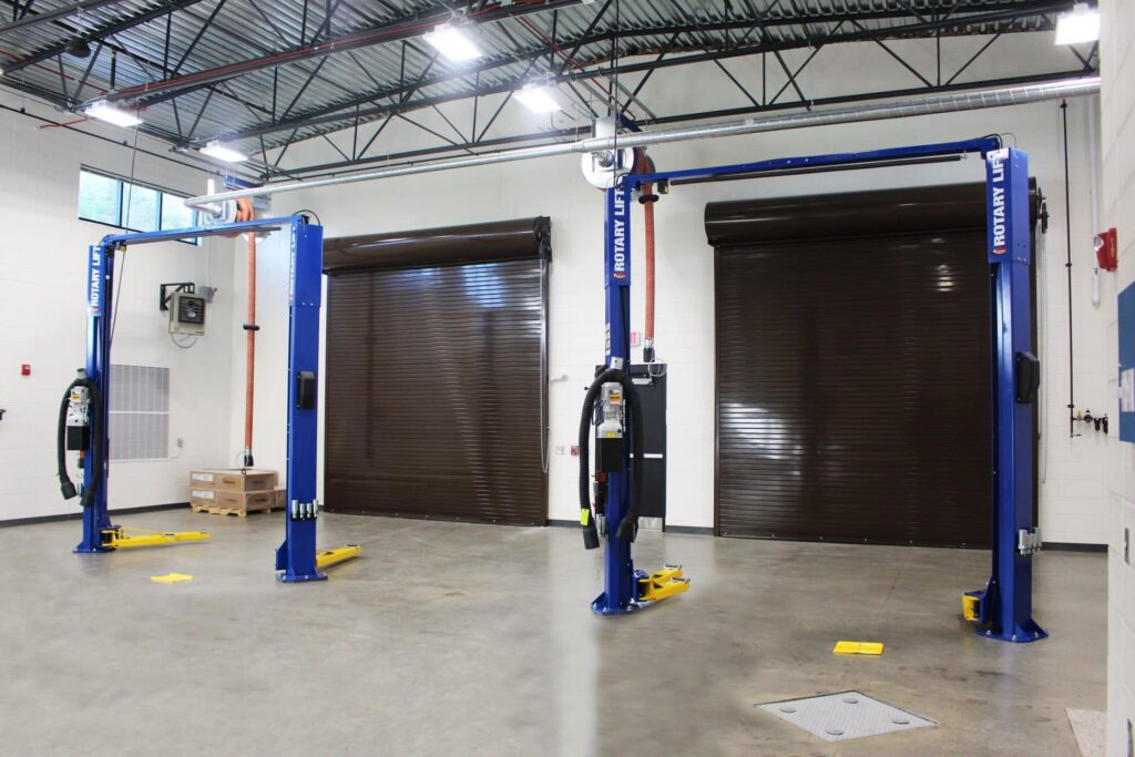 Two blue hydraulic vehicle lifts stand inside a spotless, well-lit garage featuring two large brown roll-up doors in the background and several boxes stacked along the left wall.