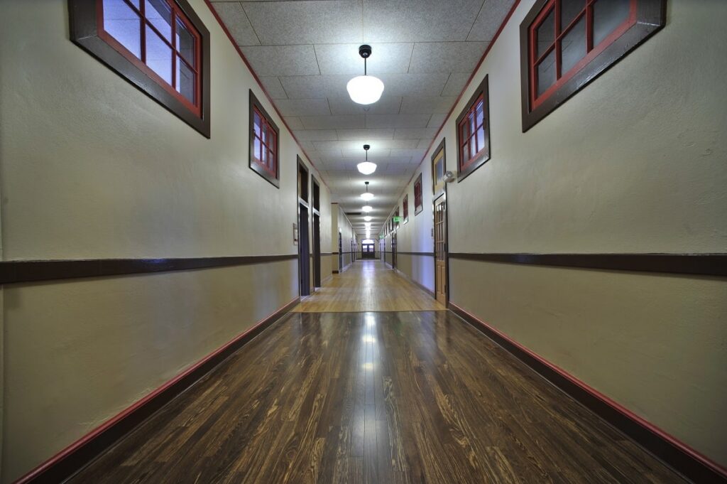 A long, empty hallway with shiny wooden floors, beige walls accented by brown trim, and small square windows positioned near the ceiling. Ceiling lights brighten the corridor as it leads toward a well-lit area at the far end.