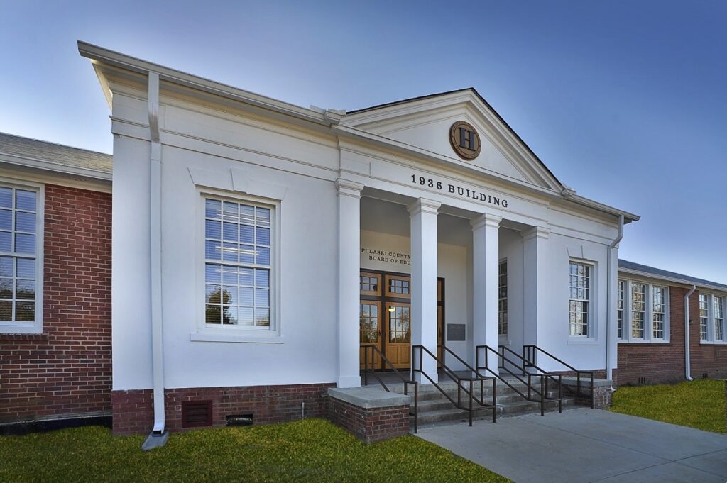 A white and red brick building labeled 1936 Building above the entrance, with steps, metal railings, large windows, and a manicured lawn under a clear blue sky.