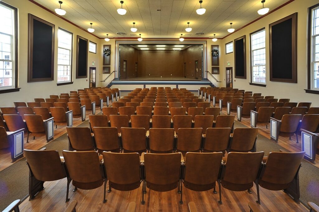 Empty auditorium featuring rows of brown wooden seats directed toward a stage, with large windows on each side, ceiling light fixtures, and glossy polished wooden floors.