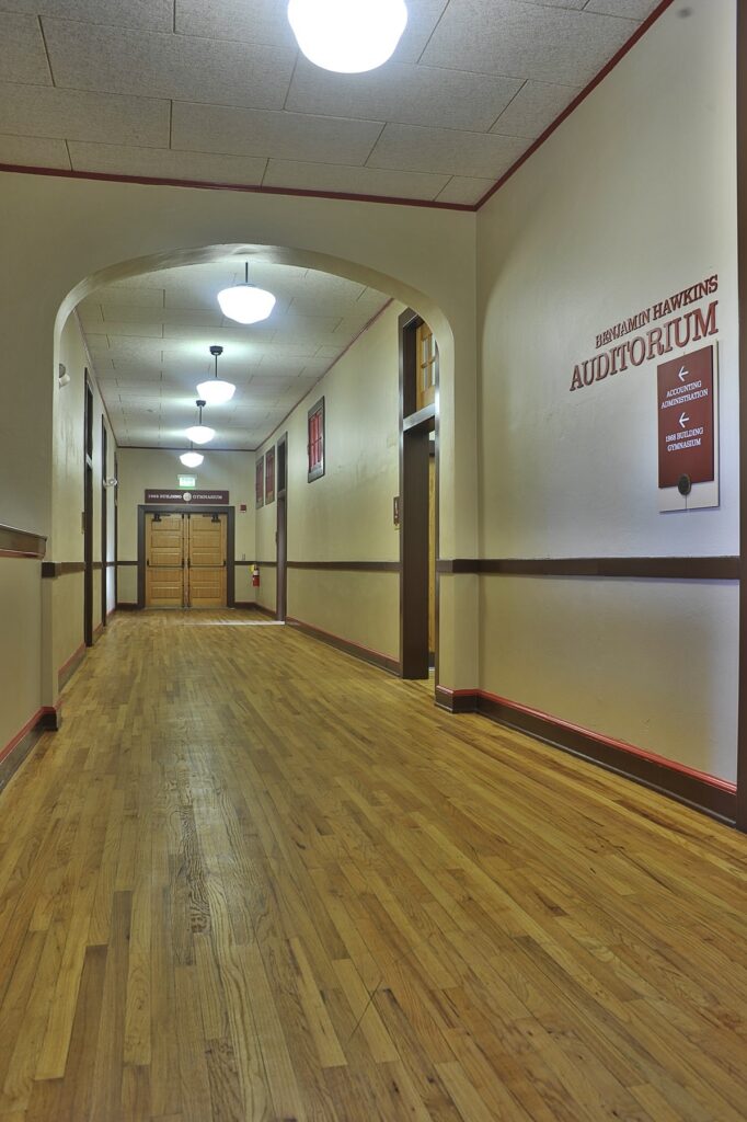 A long, empty hallway with wooden floors and beige walls leads to double doors. Signs on the right wall indicate the auditorium entrance. Ceiling lights illuminate the space.