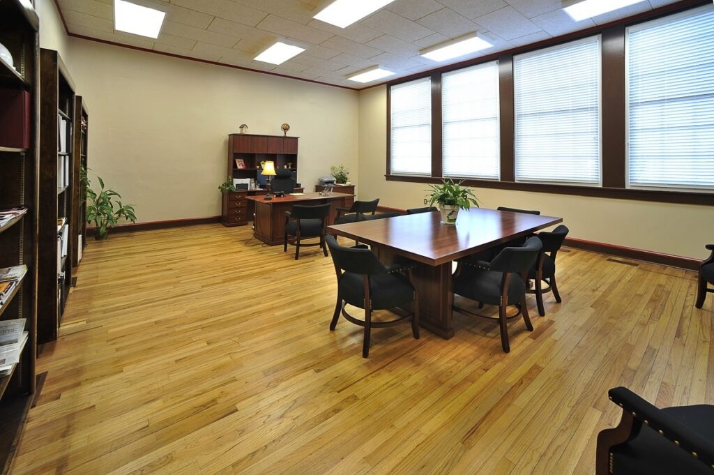 A bright, spacious office features wooden floors, a large central table encircled by chairs, bookshelves lining the walls, a desk equipped with a computer, and expansive windows that fill the room with natural light.