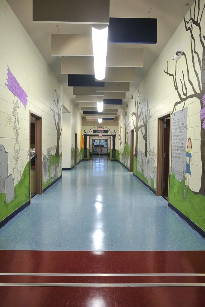 A brightly lit school hallway with murals on the walls depicting trees, gravestones, and writing. The floor is blue with a red section at the front, and doors line both sides of the hallway.