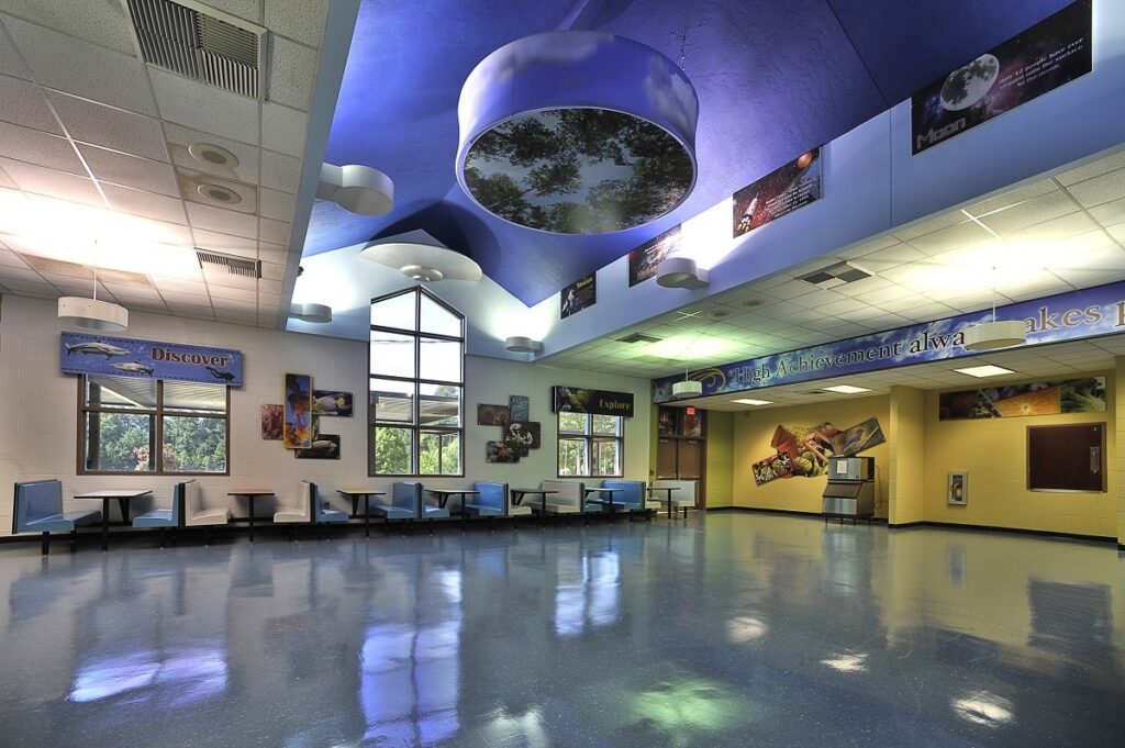 A well-lit, empty cafeteria featuring blue booths and a glossy blue floor, with large windows letting in natural light. Colorful educational posters line the walls, and the ceiling displays circular skylights along with space-themed decorations.