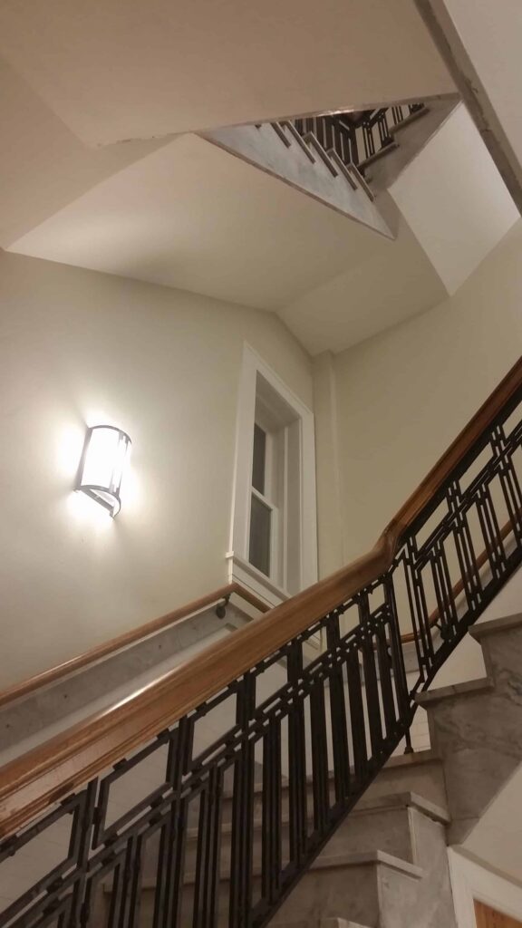 A stairwell with metal railings, a wooden handrail, and a white wall-mounted light fixture. There is a window on the landing and the view looks upward through multiple flights of stairs.
