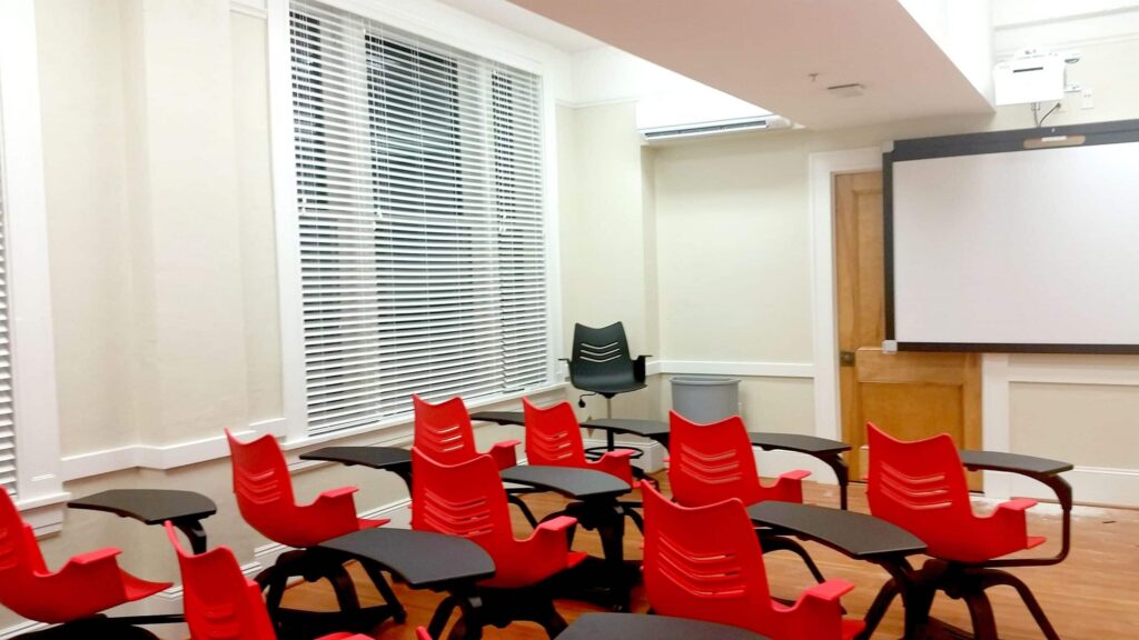 A classroom featuring red chairs and black desks arranged to face a projector screen, with large windows covered by blinds, and a single black chair positioned near a door at the back.