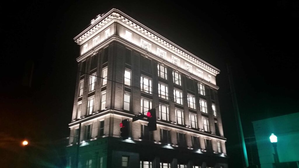 A tall, historic building glows brightly at night, its ornate white facade standing out against the dark sky. In the foreground, red traffic lights hang prominently.