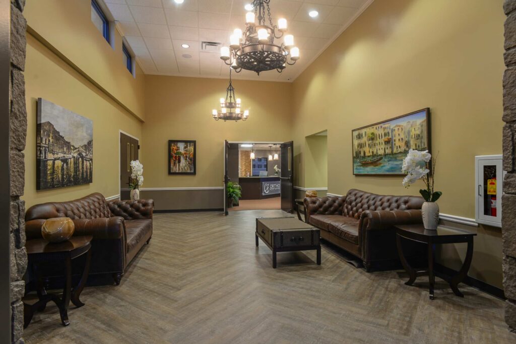 A warmly-lit lounge featuring two brown leather sofas, a coffee table, side tables adorned with white flowers, artwork on beige walls, and two chandeliers suspended from a high ceiling. An open doorway provides access to another room.