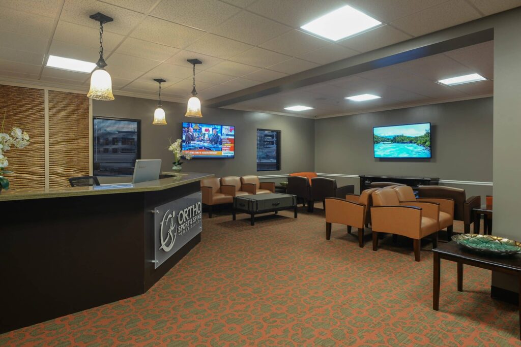A modern waiting room features tan chairs, three wall-mounted TVs, a reception desk with a sign, pendant lights, and neutral décor. The space is clean and well-lit with patterned carpet and decorative accents.